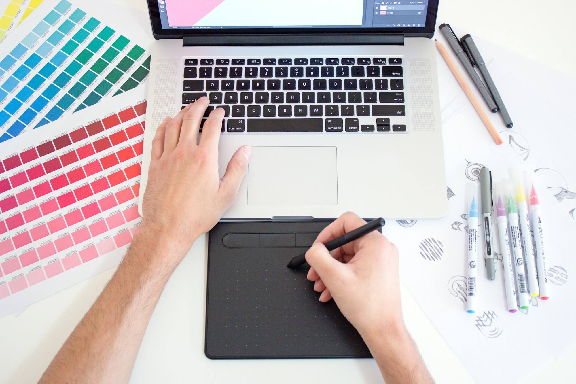 A designer working on a drawing tablet beside a laptop, colour swatches and marker pens